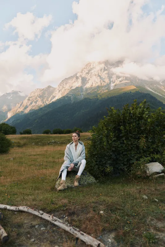 Young woman in casual clothes sitting on a rock in a mountain meadow with cloudy sky. Concept of lifestyle, nature, and outdoor relaxation in fresh air.