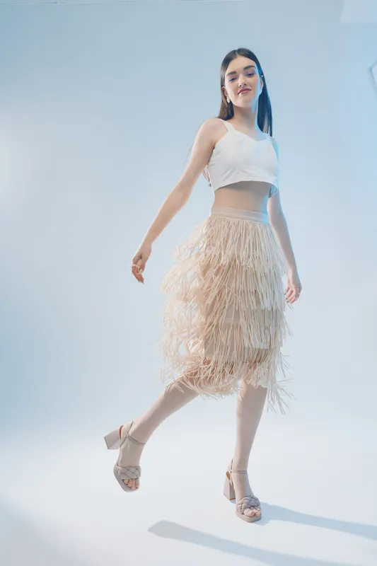 Young woman in a stylish white top and beige fringe skirt poses confidently on a light background. Concept for woman beauty fashion, elegance, and modern style.