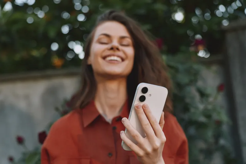 Smiling woman outdoors taking a selfie with a smartphone. Casual attire, natural light, lush garden background. Lifestyle, wellness, and self-care theme with a warm, candid mood