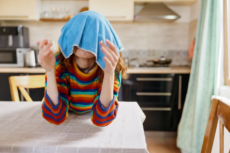 Playful girl indoors in a kitchen, with a towel on her head, rainbow sweater, candid moment, cozy home atmosphere, bright natural light, colorful fabric and scene