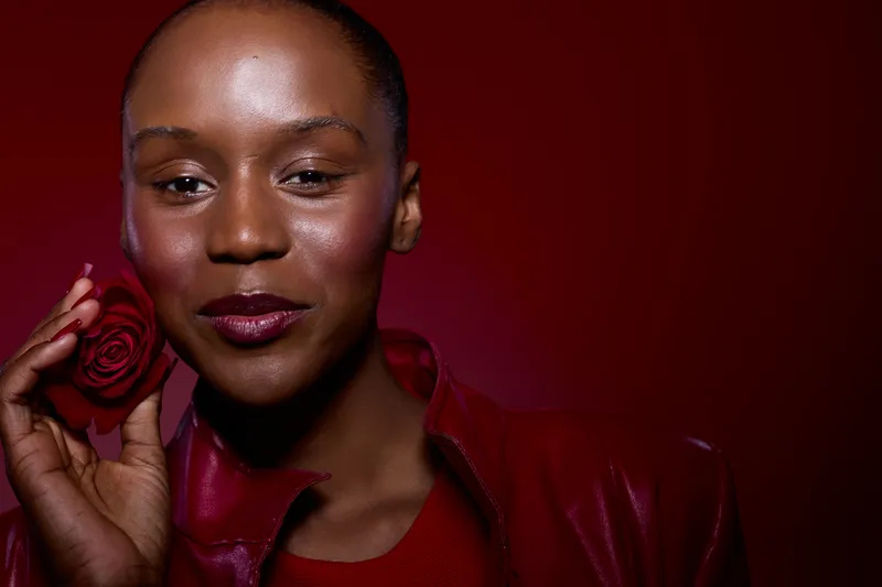 Close-up of a happy African woman with dark skin holding a red rose near her face. She wears a red leather jacket and smiles gently against a red background. Concept of beauty and elegance.