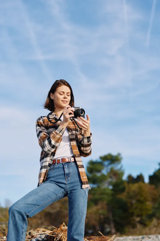 Woman photographer outdoors, plaid shirt and jeans, holding camera and aiming at scenery with clear blue sky, trees and natural landscape, capturing candid moment