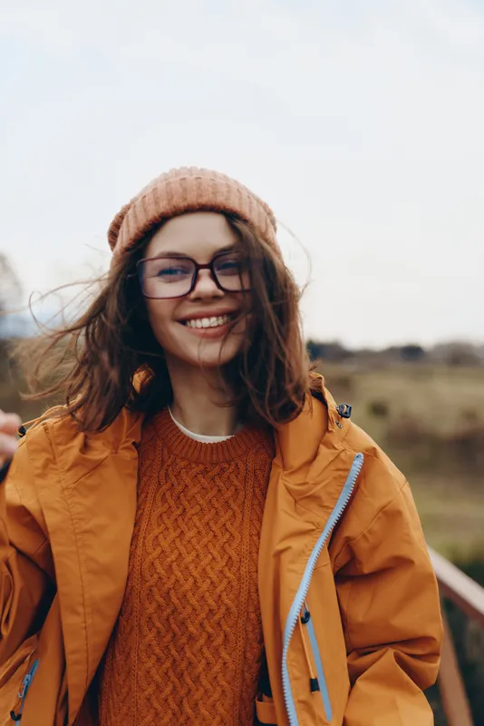 Woman lifestyle, outdoors in a rural field, smiling warmly, glasses and knit beanie, orange jacket over textured sweater, golden hour sunlight, gentle breeze, natural colors, open sky