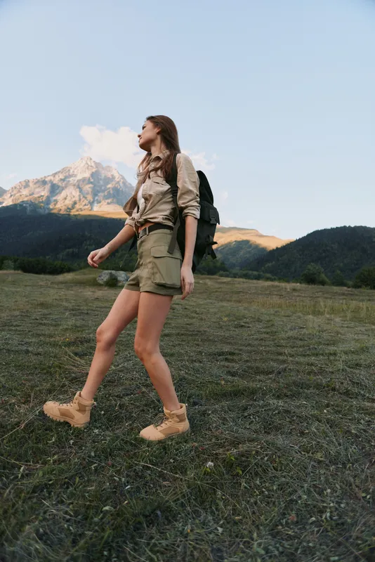 Hiking woman walking across a grassy field with a backpack, mountains in the background, daytime light. Concept of outdoor adventure, wellness, and exploration.