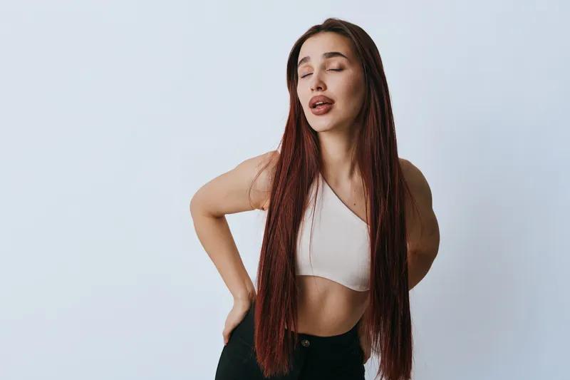 Woman with long braided hair and closed eyes holding her lower back showing discomfort or pain in casual white top and black pants against plain background.