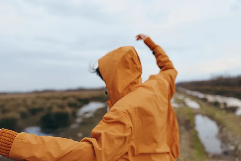 Woman lifestyle outdoors, jacket hiking, wind movement, cloudy sky, river valley landscape, orange raincoat, joyful pose, countryside trail, natural light, hood up, breezy air, now