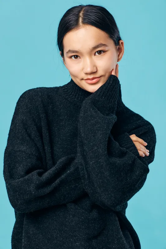 Young woman with black hair wearing a dark sweater posing with hand on face against blue background. Casual portrait, confident expression, studio shot with soft lighting.