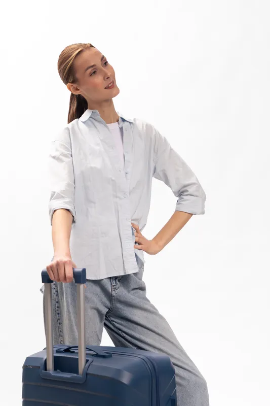 Young woman in casual outfit stands with hand on hip and holds blue suitcase handle, looking away. Concept for travel, vacation, and lifestyle with relaxed confident mood.