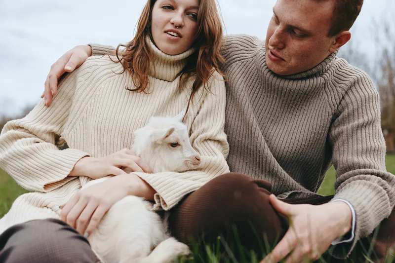 Casual couple outdoors with a small white goat kid on their lap, cozy sweaters, tender moment, natural light, rustic setting, lifestyle scene of companionship and care, warmth