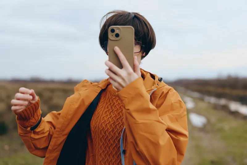 Woman outdoors taking a selfie with phone, wearing mustard jacket and knit sweater, casual lifestyle, wind lifting hair, countryside fields, soft daylight, relaxed posture