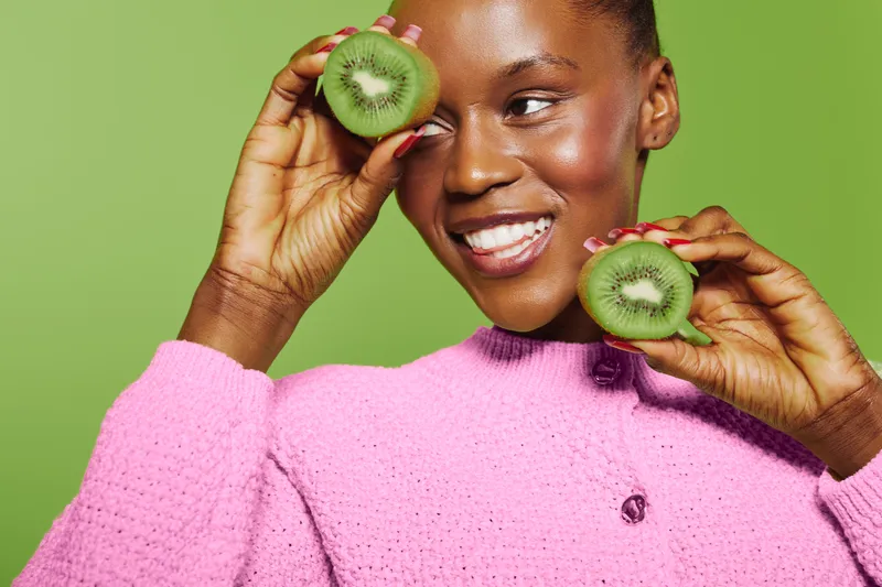 Smiling dark skinned African woman in pink sweater holds two halves of kiwi fruit near her face on green background. Concept of healthy lifestyle, natural food, and positive mood.