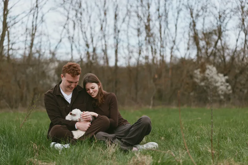 Couple sitting on the grass in a park, smiling and cuddling a small white dog, enjoying an outdoor moment together. Soft natural light and a relaxed mood create warmth and calm affection.
