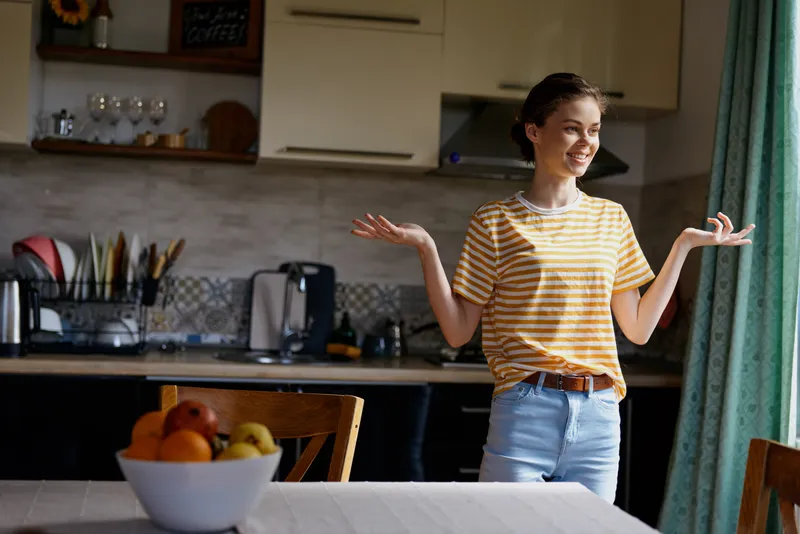 A cheerful woman stands in a bright kitchen, hands raised in a playful shrug, wearing a striped shirt and jeans. A warm, everyday home moment that conveys relaxed lifestyle and cheerful mood.