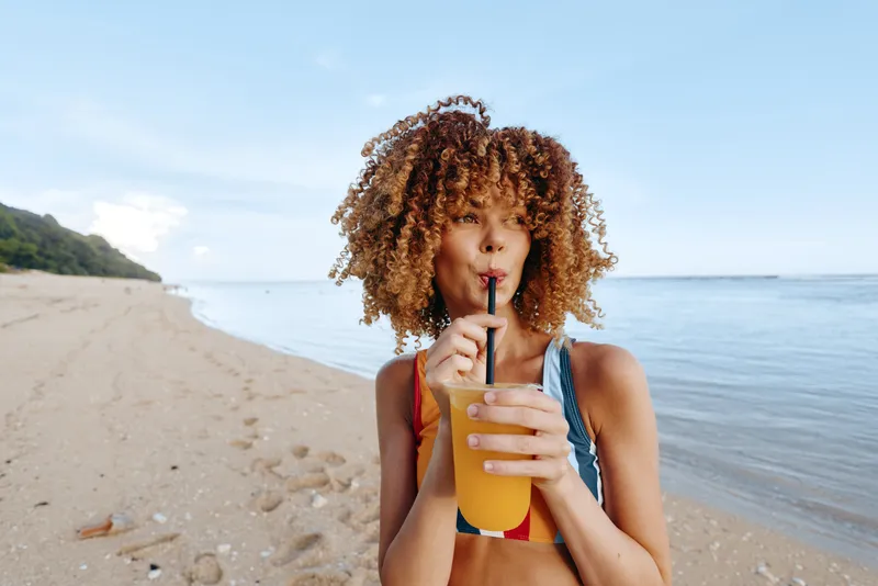 Woman with curly hair holds a refreshing drink with straw on a sandy beach, ocean horizon behind, summer vacation mood, bikini, soft light, relaxed smile