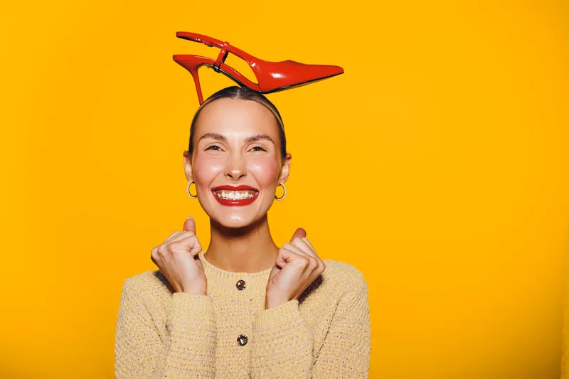 Happy woman with red lipstick and earrings smiles widely, holding red high heel shoes on her head against bright yellow background. Concept of joy, fashion, and playful mood.