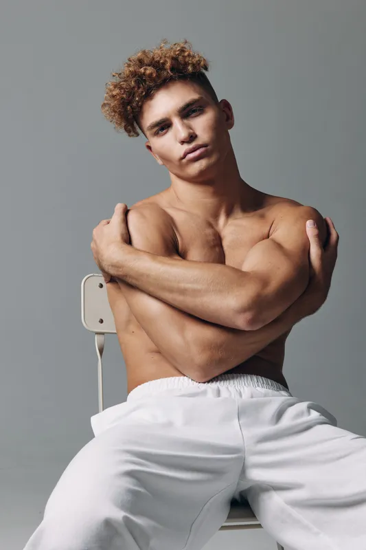 Young man with curly hair sitting shirtless on chair with arms crossed, posing in white pants against gray background in studio portrait.