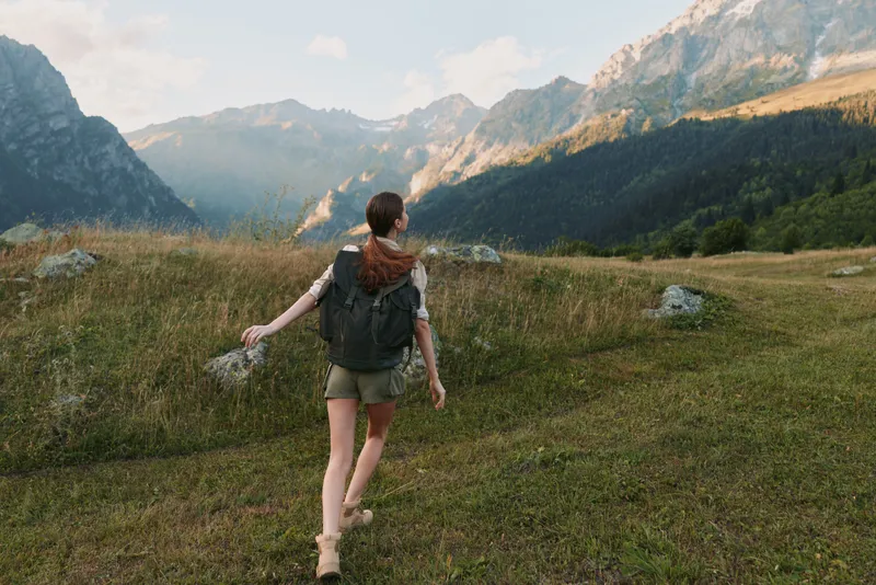 Young woman with a backpack hikes through a grassy mountain valley under a partly cloudy sky during a peaceful outdoor adventure.