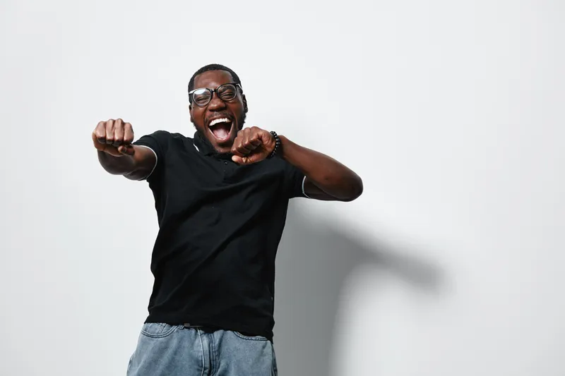 African American man with dark skin wearing glasses and black t shirt dancing and smiling with excitement on white background. Casual clothing and joyful expression in studio.