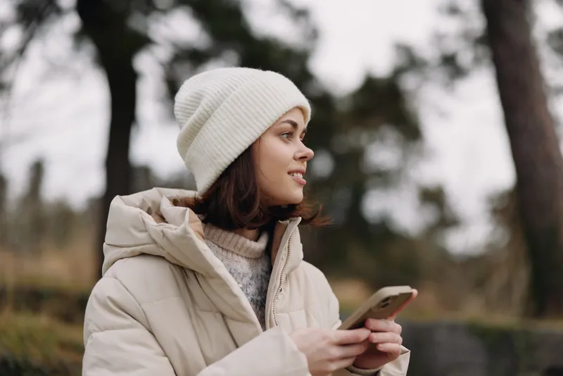 Woman in a winter coat and beanie holds a phone outdoors, a calm portrait in a park setting with trees and soft natural light, conveying a cozy and reflective mood in chilly weather.