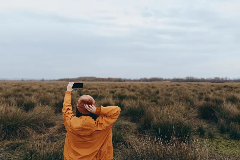 Woman lifestyle outdoors in a grassy field, wearing a hat and yellow coat, holding a smartphone to take a photo, back view under cloudy sky, peaceful nature background slow walk calm air