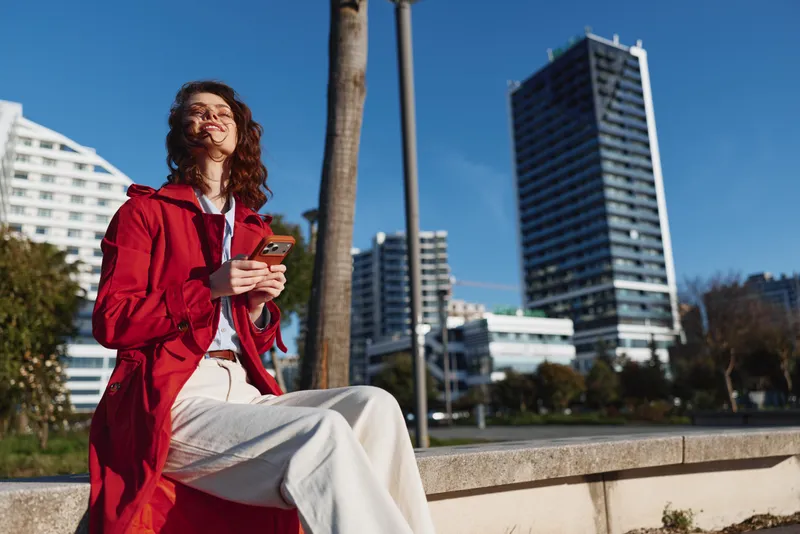 Young woman in red coat sits outdoors in city park sunny day holds smartphone smiling enjoys fresh air urban lifestyle modern buildings background.