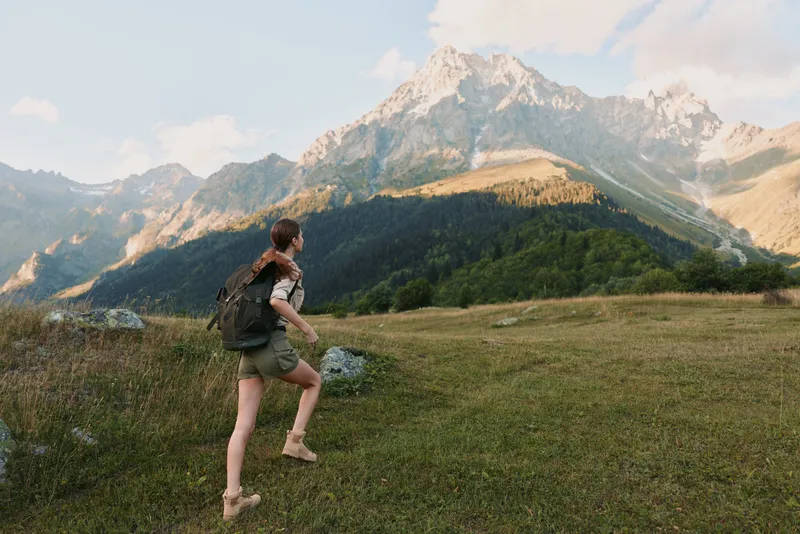 Active traveler hikes across a grassy alpine hillside toward rugged peaks, wearing a backpack, under a blue sky with distant mountains and green valleys, seeking adventure.