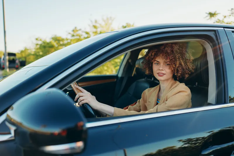 Candid woman driver in a modern car, sunlit exterior and warm retro tones, relaxed pose, natural colors, urban road backdrop, lifestyle moment captured with cinematic flair and warmth