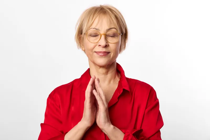 Woman prayer meditation senior hands red eyes calm studio portrait, eyes closed, hands together praying, serene expression, soft lighting, white clean background