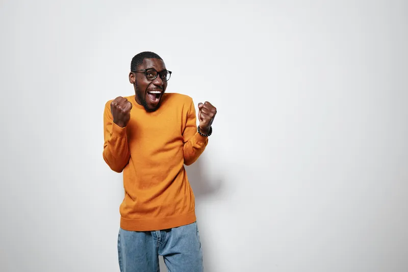 African American man with dark skin wearing orange sweater and jeans celebrating with clenched fists and excited expression on white background. Casual style and joyful mood.