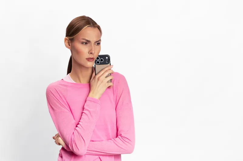 Thoughtful woman in pink sweater holds smartphone near face, looking away with serious expression. Isolated on white background. Concept for communication, decision, and technology use.