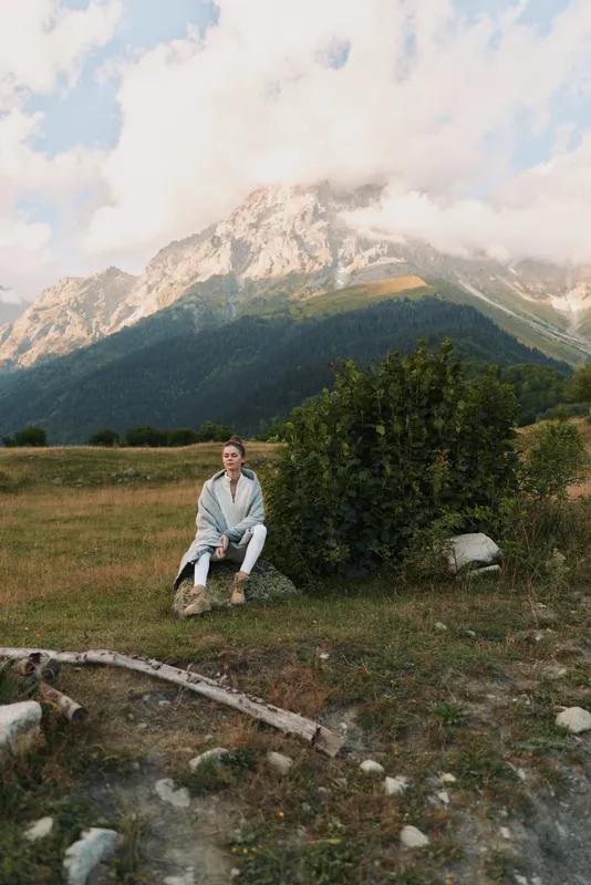 Quiet outdoor scene with a person sitting on a rock in a grassy field, mountain range in the distance, clear sky, relaxed pose, natural colors, serene lifestyle moment