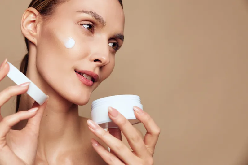 Young woman applying white face cream on her cheek with a small spatula, holding an open jar. Close-up on a beige background. Concept of skincare, beauty, and self-care routine.