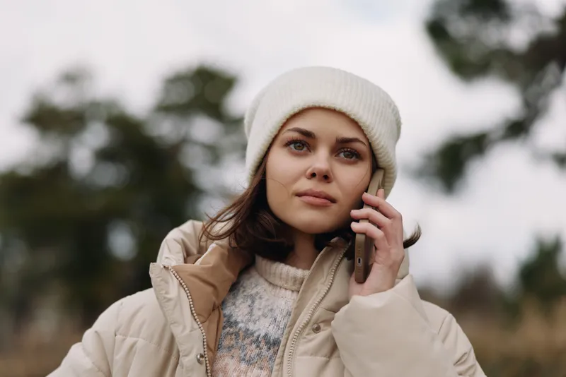 Woman wearing a winter coat and beanie talks on the phone outdoors, calm expression, soft natural light, casual portrait in chilly weather with blurred trees in the background.