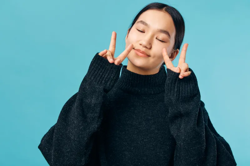 Young woman in black sweater with eyes closed smiling and showing peace signs with both hands on blue background. Casual portrait, relaxed, gesture and happiness concept.