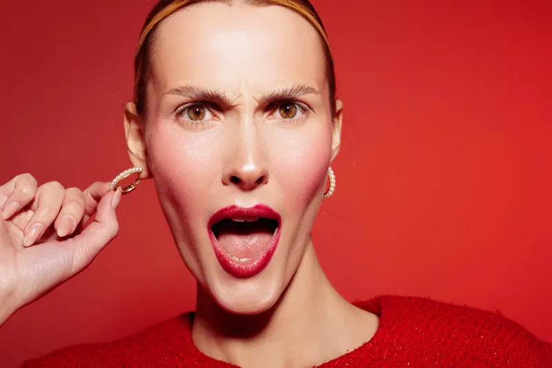 Close-up of a surprised woman with red lipstick and earrings pulling her ear against a red background. Concept for emotion, expression, beauty, and fashion portrait.