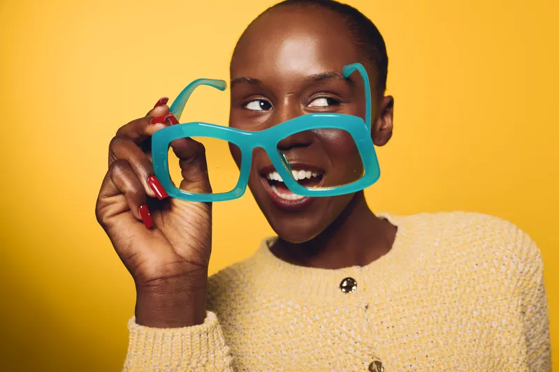 Smiling dark skinned African woman holds oversized blue glasses near face on yellow background. Concept of joy, fashion, playful mood, and creative portrait of black female model.