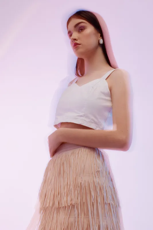 Young woman in white top and beige fringed skirt poses confidently against soft gradient background. Concept of woman beauty fashion and modern style in studio lighting.