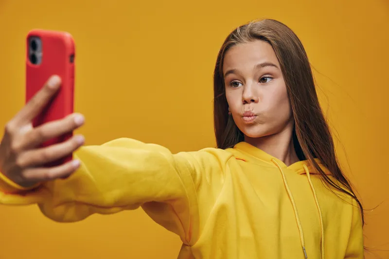 Girl in yellow hoodie taking selfie with red smartphone against orange background. Young female holding mobile phone at arm’s length making funny face for photo.