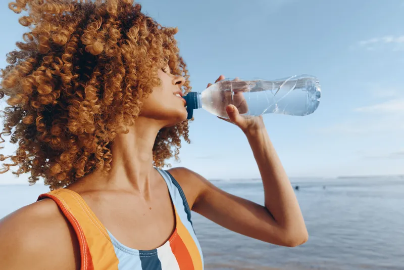 Energetic woman with curly hair drinks water from a bottle at the beach, showcasing hydration and fitness in a sunny seaside scene, casual summer attire, outdoor activity by the ocean