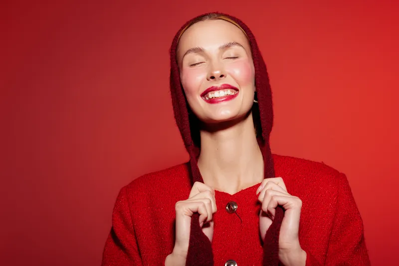 Smiling woman in red sweater and hood stands against red background with closed eyes and bright lipstick. Concept of happiness, fashion, beauty, and vibrant color contrast in studio.