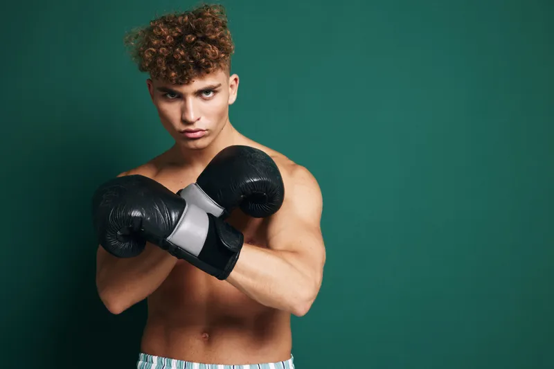 Boxer man with curly hair posing with gloves crossed, focused and ready for fight or workout against green studio background with muscular body and intense look.