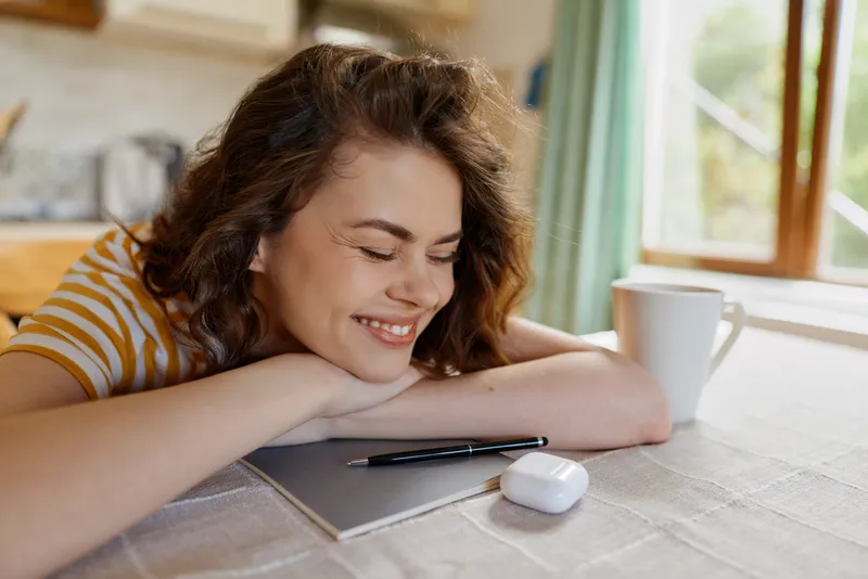 Smiling woman rests her head on a desk beside a notebook and pen, in a sunny home setting. Warm, inviting mood suggests relaxed daily routine and casual creativity.
