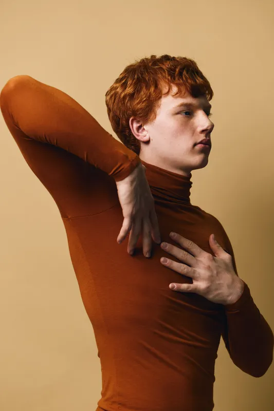 Young man with red hair wearing brown turtleneck posing with one hand on chest and the other arm raised against beige background in studio portrait.