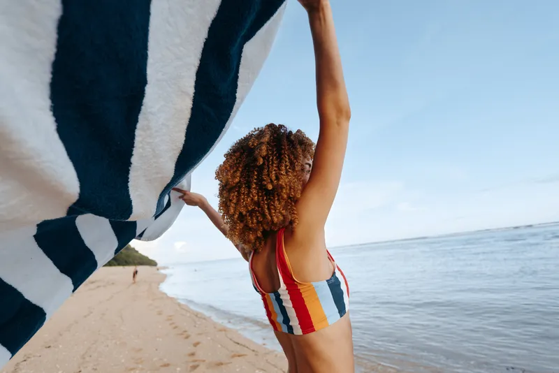 Beach ocean yoga stretching woman swimsuit sunlight summer balance pose at seaside, athletic female stretches arms overhead on sand, towel on shoulder, braided hair, calm blue horizon, vacation