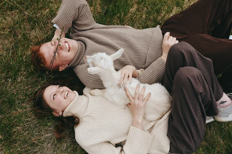 Young couple lying on grass in cozy sweaters happily plays with a white baby goat enjoying a relaxed lifestyle moment outdoors in casual autumn clothing.
