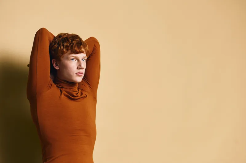 Young man with red hair in brown turtleneck poses with arms behind head against beige background. Portrait, casual style in warm tones with thoughtful expression.