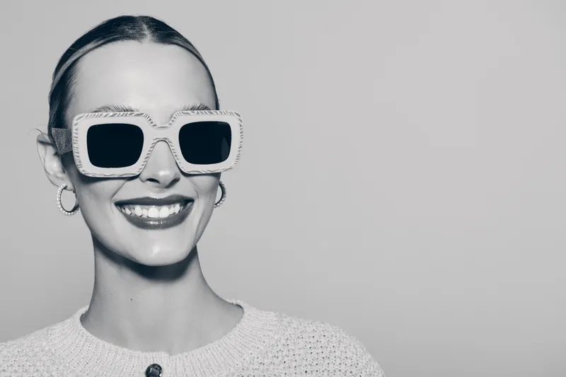 Smiling young white woman with fair skin wearing oversized sunglasses and a knitted sweater, isolated on a neutral grey background.