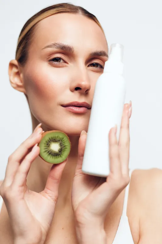 A young woman with clear skin holds a white cosmetic bottle and a sliced kiwi fruit close to her face, symbolizing natural skincare and healthy beauty routine concept.