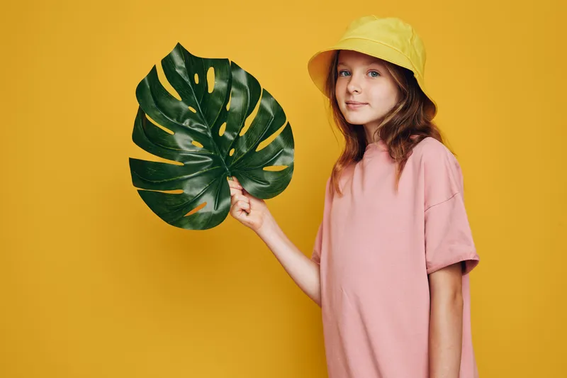 Girl in a pink shirt and yellow hat holding a large green tropical leaf on a yellow background. Studio portrait with bright colors and natural expression.