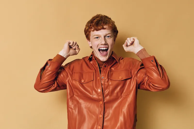 Young man with red hair wearing brown leather jacket celebrates with fists raised and smiling excitedly on beige background. Portrait of happy male expressing joy and success.